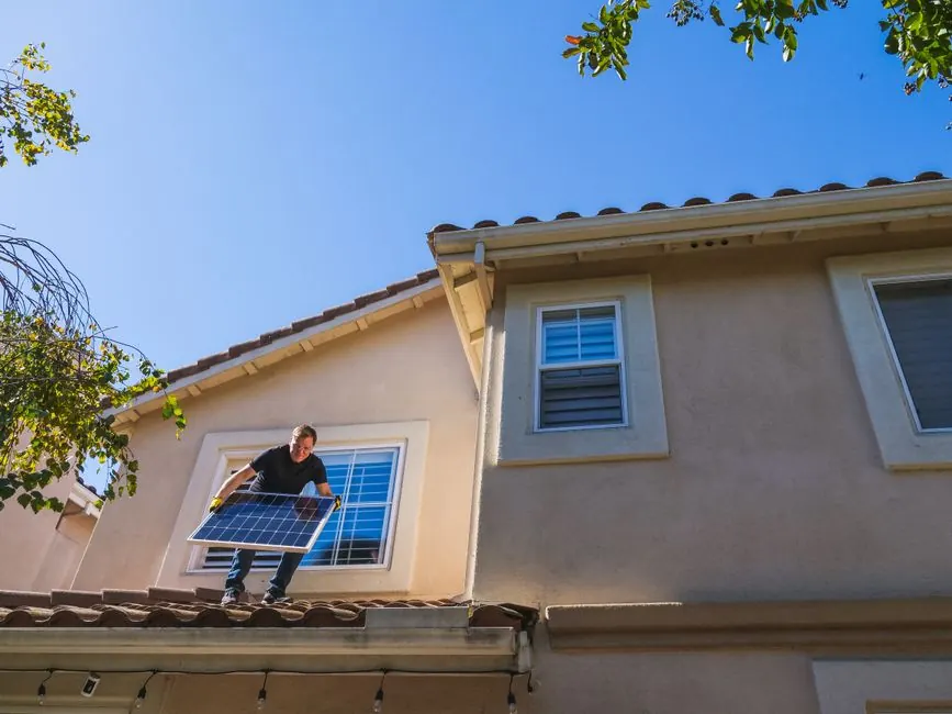 Man installing a solar panel on a rooftop under a clear blue sky, promoting renewable energy.